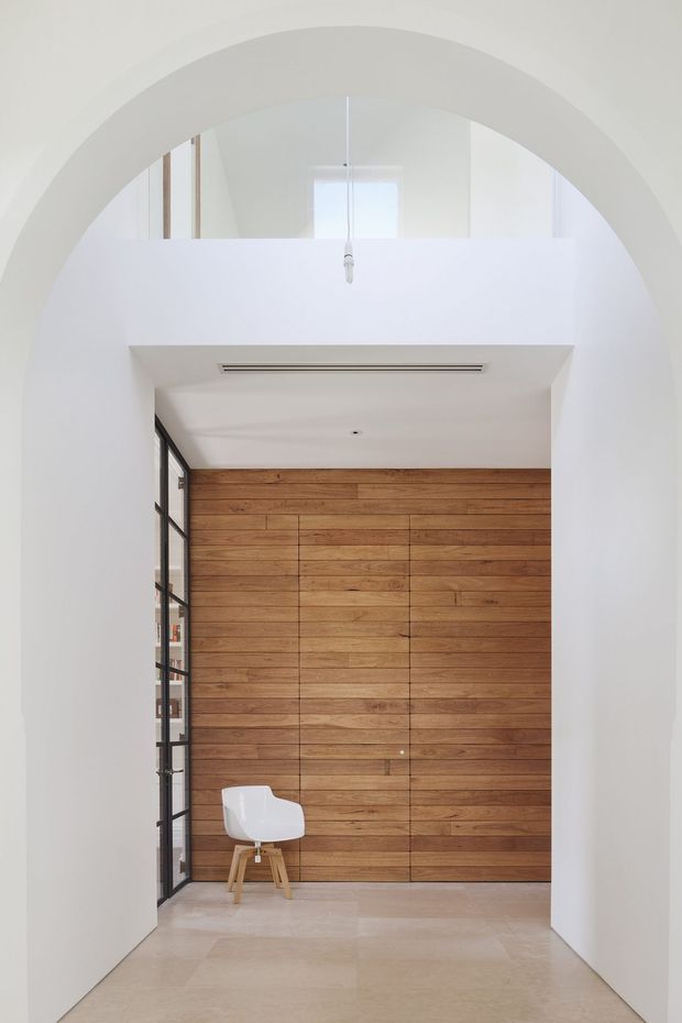 A minimalist interior entryway or hallway framed by a large white arched opening. The space features smooth white walls and ceiling, a pale polished stone floor, a full-height warm timber-panelled feature wall with concealed joinery, and a black-framed glazed door or partition along the left side. A single white chair provides the only loose furnishing. The tall opening and upper glazing create an airy, light-filled sense of vertical space.