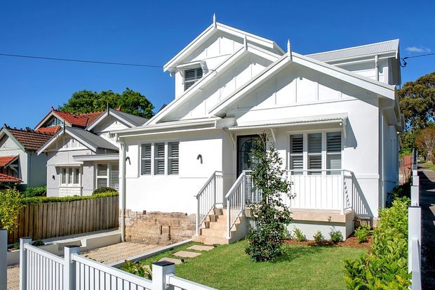 Bright exterior view of a renovated white timber-clad house with prominent front gables, decorative verge and bargeboard detailing, louvred windows, a raised entry porch, steps, and a small landscaped front garden. Neighbouring houses are visible in the background.