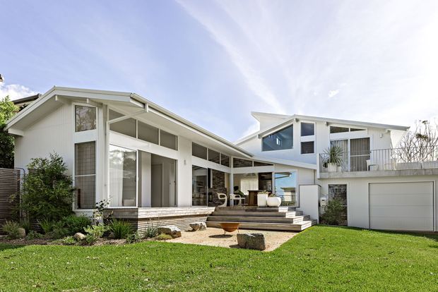 Exterior view of a bright, low-rise modern coastal-style home with white weatherboard cladding, broad glazed openings, angled pitched rooflines, and a stepped terrace leading to a landscaped lawn. A covered outdoor sitting area and attached double garage are visible.
