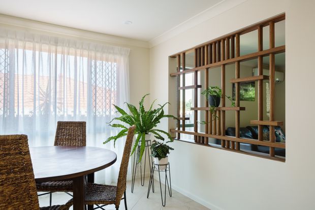 Bright dining area with a round dark timber dining table and woven rattan chairs. Sheer curtains provide filtered daylight, while a large timber decorative screen with open shelving separates the room from an adjacent space. Indoor ferns and potted plants add a natural accent.