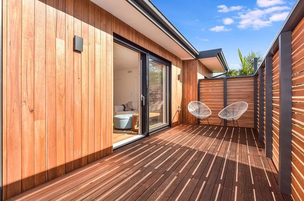 A sunny timber-clad outdoor deck or terrace enclosed by horizontal timber privacy screens and fencing. The deck features dark timber decking, a sliding glazed door opening into an interior room, and two wire-frame lounge chairs. The low-pitched roof and warm wood surfaces create a contemporary residential outdoor space.