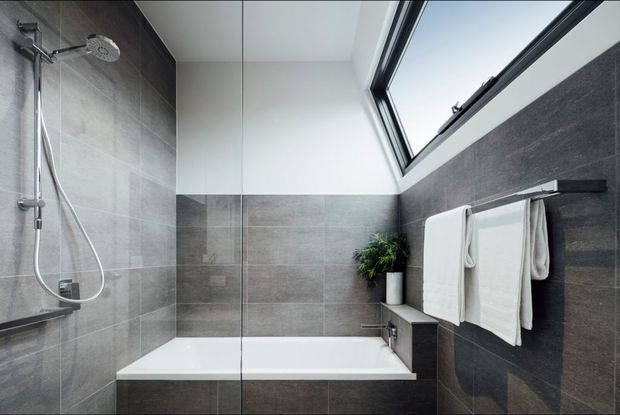 Contemporary bathroom featuring a frameless glass shower screen, wall-mounted shower fittings, a white bathtub, dark grey large-format wall tiles, a white upper wall and ceiling, a skylight, and a simple towel rail with folded white towels. A small potted plant adds greenery near the bath.