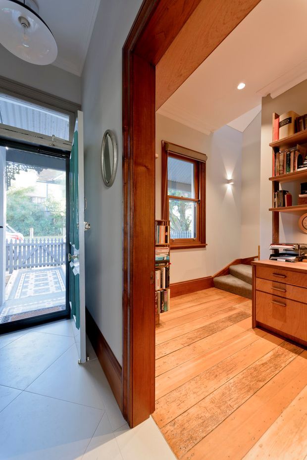 View from an entryway into a warm timber-lined study or home office. The foreground has pale floor tiles, a glazed exterior door, dark stained timber trim, and a round wall mirror. Beyond the wide timber-framed opening is a room with honey-toned timber floorboards, built-in timber bookshelves and desk cabinetry, a window, recessed lighting, and a short set of carpeted steps. The interior has traditional detailing and strong natural timber character.