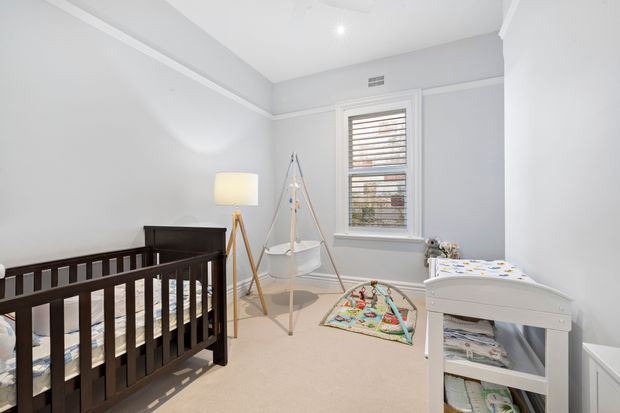Bright, softly styled nursery with a dark timber cot, white changing table, hanging baby bassinet, floor lamp, play mat, and a shuttered window. The room has pale grey walls, white trim and ceiling, and beige carpet, creating a calm, practical atmosphere.