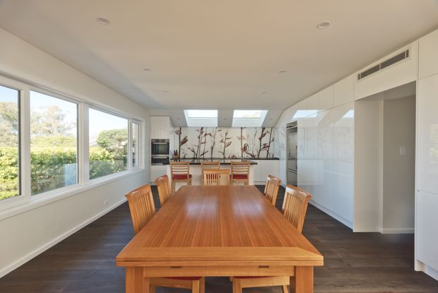 Bright open-plan dining area with a long solid timber dining table and matching chairs, large side windows overlooking greenery, dark timber flooring, and extensive glossy white built-in cabinetry along the right wall. A skylit rear section includes decorative branch-patterned wall artwork or a feature panel, creating an airy contemporary family dining space.