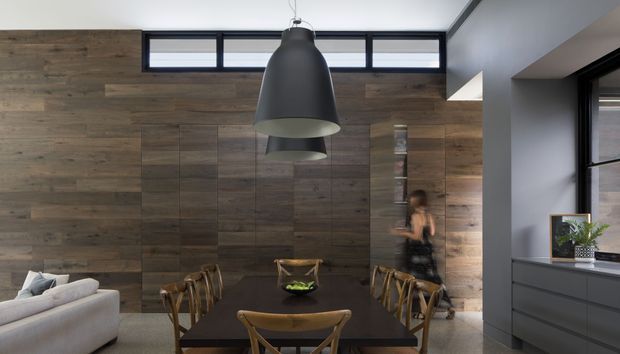 Contemporary dining area with a long dark dining table and timber chairs, backed by a full-height feature wall of varied dark timber panels. A large black pendant light hangs above the table, with clerestory windows providing natural light. Part of a light sofa is visible at left, while dark built-in cabinetry and a window appear at right.