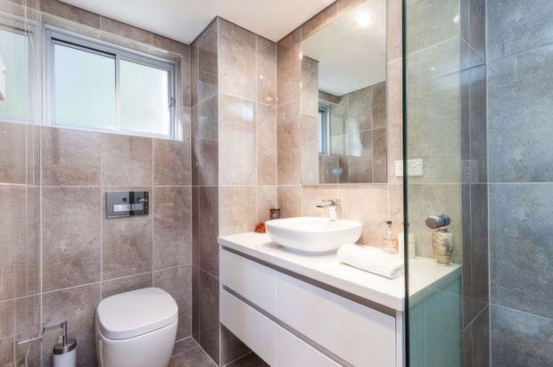 Modern bathroom with large-format taupe and grey wall and floor tiles, a wall-hung white vanity with basin, concealed cistern toilet, frameless glass shower screen, and large mirror. Natural light enters through a frosted window.