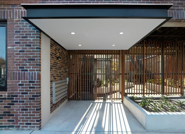 A contemporary covered entryway or carport framed by dark red-brown brickwork. The flat projecting canopy has a white soffit with recessed downlights, while vertical timber-look slatted screens create privacy and patterned shadows across the concrete paving. A raised planter bed with greenery sits to the right, and a glazed entry area is visible behind the screen.