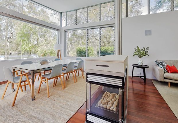 Bright, airy open-plan interior with a dining area and adjacent living space. Floor-to-ceiling corner glazing provides abundant natural light and views of trees and water outside. The room features a light timber dining table with upholstered chairs, a pale upholstered sofa, a compact black side table, a large neutral woven rug, dark polished timber flooring, and a freestanding fireplace or heater cabinet in the foreground.