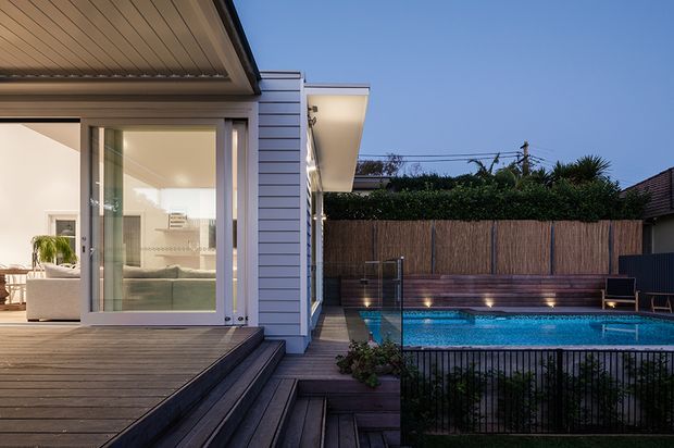 Dusk exterior view of a contemporary single-storey home opening onto a timber deck and raised swimming pool. The house features white horizontal cladding, broad sliding glass doors, a covered soffit, and warm interior lighting. The pool terrace includes timber privacy screening, integrated step lighting, planting, and black safety fencing.
