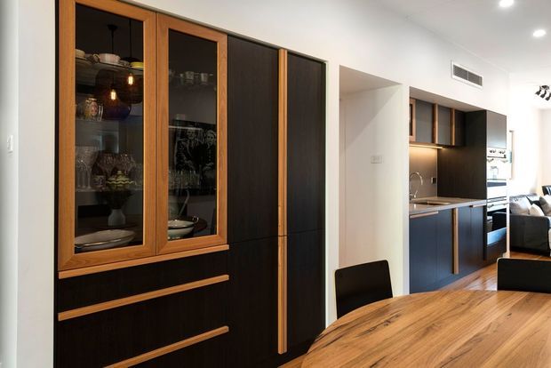Contemporary open-plan dining and kitchen interior featuring a large timber dining table, dark integrated cabinetry with warm timber detailing, glass-front display cabinets, a compact kitchen with under-cabinet lighting, and a partially visible living area beyond.