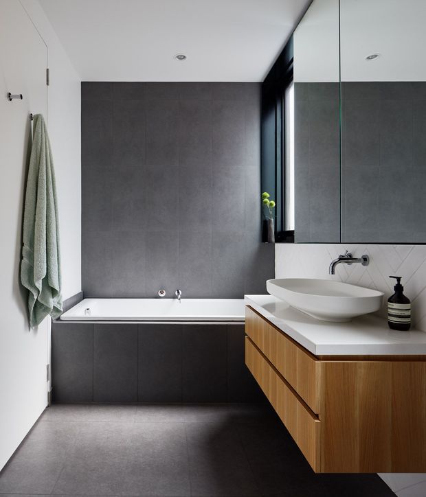 A compact contemporary bathroom featuring a built-in white bathtub, large dark grey wall and floor tiles, a floating timber vanity with a white countertop and sculptural vessel basin, wall-mounted chrome tapware, mirrored upper cabinetry, and a narrow black-framed window. The room has a clean, restrained palette with warm timber contrast and recessed ceiling lighting.