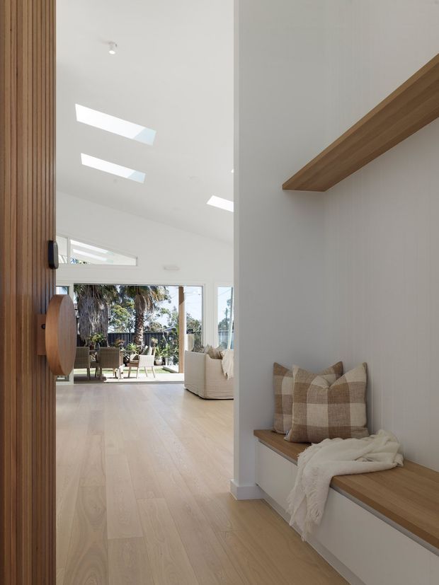 Bright contemporary residential entryway or hallway with a full-height vertical timber-slatted door on the left, pale timber flooring, white walls, and a built-in timber bench with cushions and a throw. The hallway opens into a light-filled living area with large glazing, skylights, and an outdoor view. A floating timber shelf adds architectural warmth above the bench.
