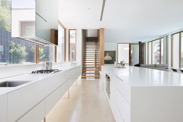 Bright, spacious contemporary open-plan kitchen with extensive white handleless cabinetry and a large white island. The room features pale polished flooring, a linear gas cooktop, integrated appliances, full-height glazing along one side, and a timber-slat stair screen leading toward the living area. The restrained palette and abundant daylight create a clean, airy atmosphere.