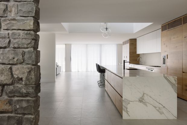A bright, open-plan contemporary kitchen viewed from the living area. The space features a long stone island with integrated seating, light timber cabinetry, white overhead cupboards, a marble-look benchtop, pale tiled flooring, full-height sheer glazing, a skylight, and a prominent rough stone masonry column in the foreground.