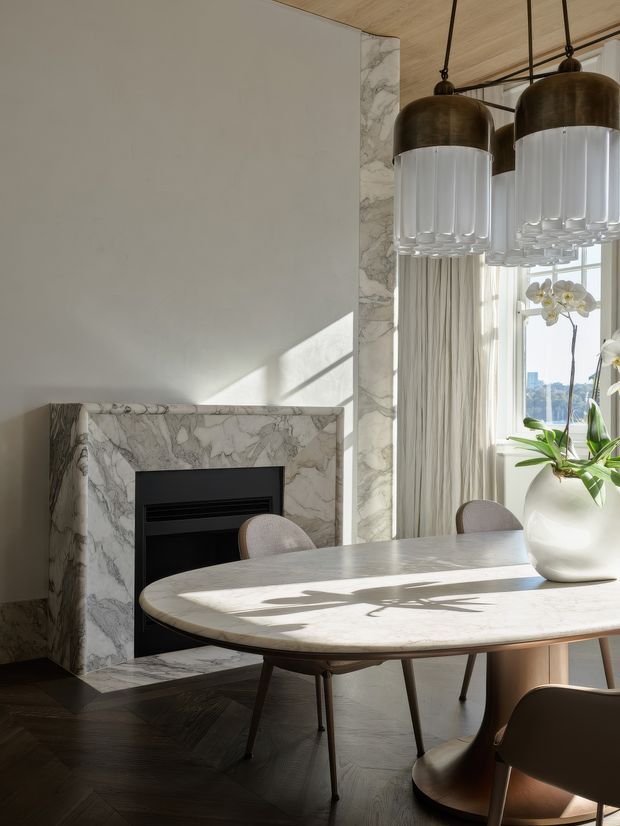 Sunlit dining area with a sculptural oval marble dining table, upholstered dining chairs, a marble fireplace surround, dark herringbone timber flooring, sheer curtains, and large statement pendant lights with bronze-toned domes and ribbed glass shades. A potted orchid sits near the window, with water and city views visible beyond.
