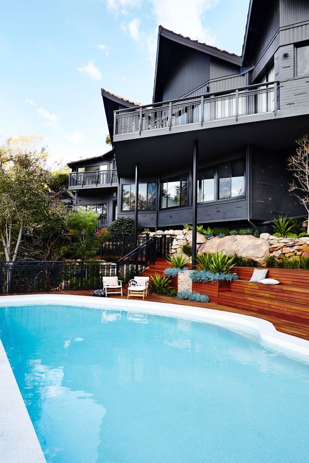 Exterior view of a multi-level dark-clad residence overlooking a bright swimming pool. The house features vertically ribbed dark cladding, pitched tiled roof sections, extensive balconies with metal balustrades, large windows, and stepped timber pool decking integrated into a landscaped sloping site with stone retaining walls and ornamental planting.