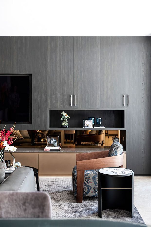 A refined contemporary living room featuring a full-height dark grey wood-grain built-in media wall with concealed storage, open shelving, and a mounted television. A warm taupe built-in cabinet and reflective backsplash sit below, accompanied by upholstered lounge chairs, a pale sofa, patterned area rug, and a small black-and-white side table. The room has a sophisticated, moody palette with warm timber and metallic accents.
