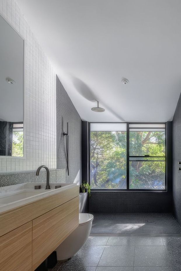 A bright contemporary bathroom with a floating light-wood vanity, integrated basin and dark curved metal tap. The walls combine small white square tiles around the vanity with dark textured tiles in the shower and window area. A freestanding-style toilet, wall-mounted shower, large mirror, and black-framed windows overlooking greenery complete the space. Grey tiled flooring and a clean, refined palette create a calm modern atmosphere.