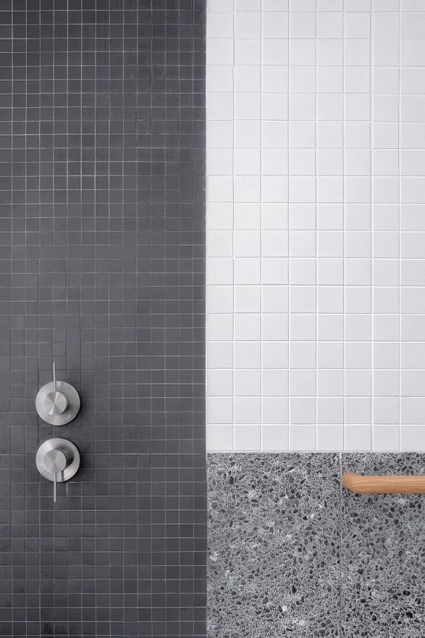 Close-up detail of a tiled bathroom or wet-area wall, split between dark charcoal square tiles and white square tiles. Two circular brushed-metal controls are mounted on the dark tiled section, while a grey speckled stone or terrazzo-like surface and a partial timber-handled element appear along the lower right edge. The composition is minimalist and material-focused.