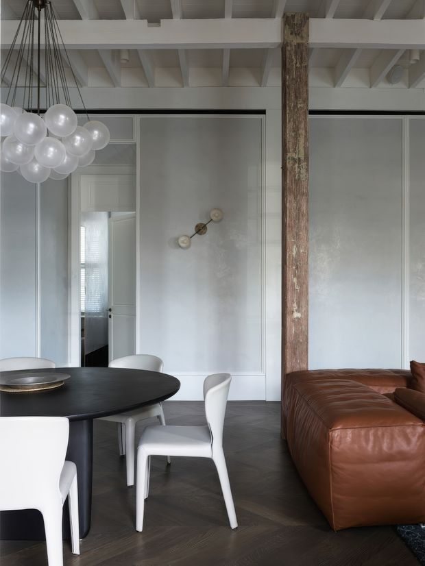 Refined open-plan dining and living interior with a dark round dining table, white sculptural dining chairs, a large brown leather sectional sofa, pale grey panelled walls, and exposed white-painted ceiling beams. A weathered timber structural post divides the dining and lounge areas, while a clustered globe pendant light hangs above the table. Dark herringbone timber flooring and restrained contemporary furnishings create a polished, slightly moody atmosphere.