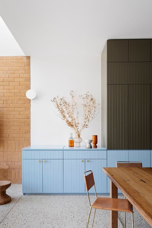 A bright contemporary dining and living space featuring a pale blue fluted built-in sideboard, dark vertically panelled cabinetry, a warm exposed brick wall, terrazzo flooring, and a solid timber dining table with leather-and-metal chairs. Minimal decorative vessels and dried branches sit on the sideboard, while a simple white wall light adds a soft accent.