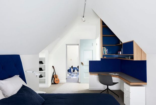 A bright attic-style bedroom with a sloped white ceiling, deep blue upholstered bedhead and bedding, and a built-in blue-and-timber study nook. The study area includes open shelving, a timber worktop, white drawer units, and a grey office chair. White built-in shelving and a partially open door lead to a bright adjoining space.