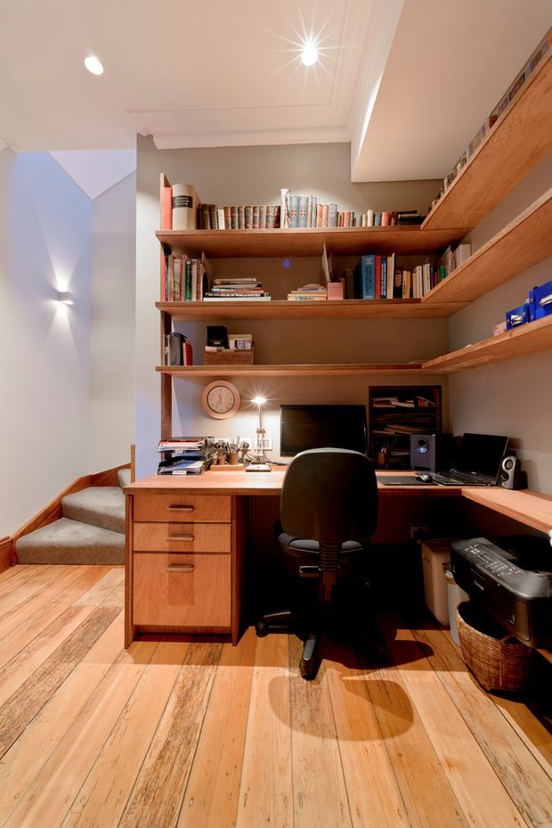 A compact built-in home office occupies a nook beside a short staircase. Warm timber flooring, a timber desk with integrated drawers, wraparound timber bookshelves, a computer workstation, office chair, and desk accessories define the space. The room has warm grey walls, white recessed and coved ceiling detailing, and layered lighting, creating a practical, warm study atmosphere.