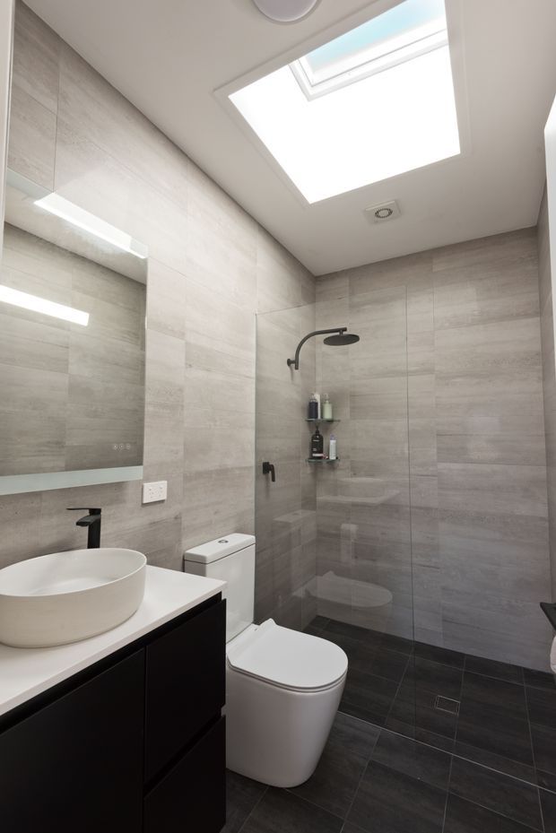 A compact contemporary bathroom with a wall-to-wall glass shower enclosure, rain shower, wall-mounted controls and recessed shelving. Large light grey stone-look tiles cover the walls, while dark grey floor tiles create contrast. A black vanity with a white vessel basin and black tap sits beside a white toilet. A large skylight provides abundant natural light, with a mirrored wall cabinet and recessed lighting adding functionality.