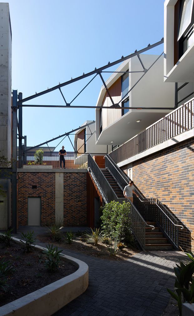 A sunlit multi-level urban residential courtyard with dark brick walls, extensive external stairs and black metal balustrades. White contemporary apartment façades with projecting balconies and timber-toned vertical cladding rise above the courtyard. A large black steel overhead structure spans between buildings, while curved concrete garden edging and low drought-tolerant planting define the paved walkway.