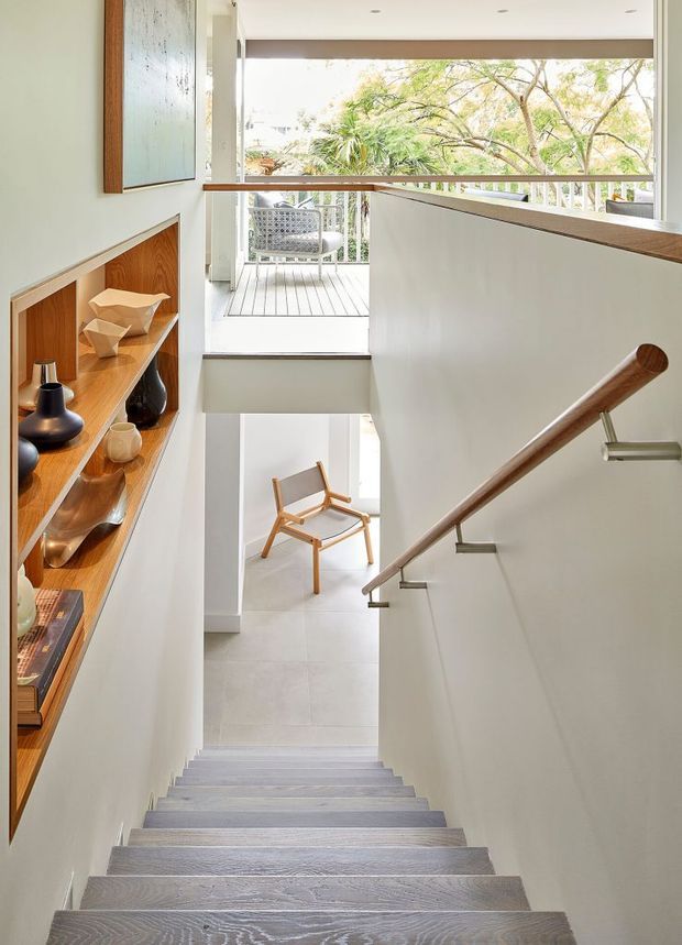 Bright, minimalist interior view looking down a straight staircase. The stair treads have a pale grey-washed timber finish, with a simple timber handrail on slim metal brackets and white walls. Built-in open timber shelving displays ceramics and decorative objects along the stair wall. At the lower level, a light chair sits near a large opening to a small outdoor balcony or terrace with leafy vegetation beyond.