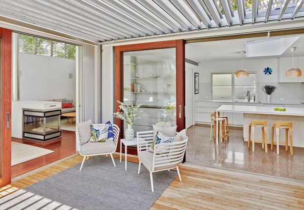 Bright open-plan indoor-outdoor living area with a contemporary white kitchen, large island, timber-framed glazed doors, and a covered deck furnished with modern outdoor lounge chairs. A timber-lined adjacent room with a desk is visible to the left, while overhead operable louvres or pergola slats create a sheltered connection to the exterior.