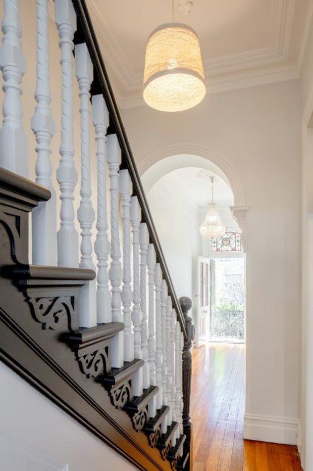 A bright, narrow traditional hallway featuring an ornate staircase with white turned balusters, dark stained timber handrails and decorative carved stair brackets. The corridor has polished timber flooring, white painted walls, high decorative ceiling mouldings, an arched opening, and pendant chandeliers leading toward a glazed entry door.