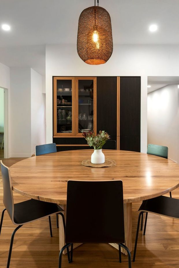A contemporary dining room with a large round solid timber dining table surrounded by dark-framed chairs. A black timber built-in cabinet with glass-front display doors and open shelving occupies the rear wall. The room has white walls and ceiling, timber flooring, recessed ceiling lights, and a prominent woven pendant light above the table. A vase of flowers forms a simple centrepiece.