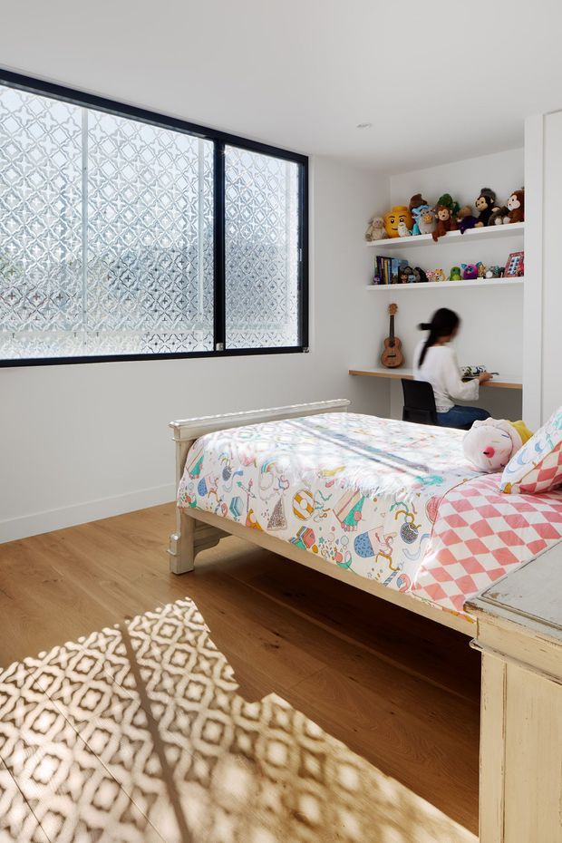 Bright children's bedroom with a single bed dressed in colourful patterned bedding, a built-in desk and chair, open display shelves filled with toys and books, and a guitar. The room has white walls, warm timber flooring, dark-framed patterned glazing, and a soft patterned rug.