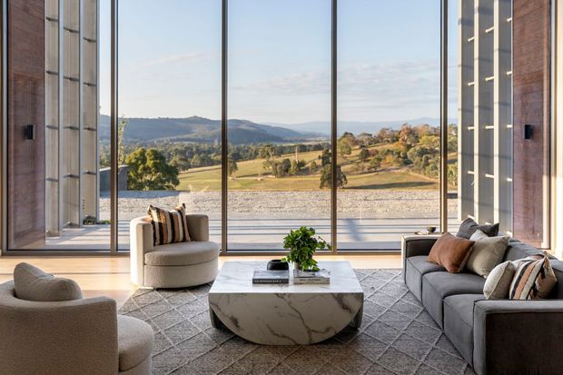 Contemporary living room with expansive floor-to-ceiling glazing framing a rural valley and rolling hills. The room features pale timber flooring, a large grey patterned rug, a curved marble coffee table, a grey sofa with neutral and rust-toned cushions, rounded upholstered lounge chairs, and tall vertical timber or wood-toned wall panels. The composition is bright, refined, and strongly connected to the landscape.