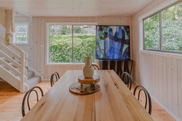 A bright timber-lined dining area with a long natural wood dining table and curved dark metal chairs. A compact staircase rises along the left wall, while large windows provide views of greenery. Pale timber wall and ceiling panelling, light wood flooring, and an abstract artwork create a warm contemporary cabin-like interior.