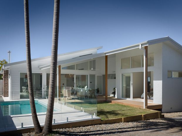 A bright single-storey modern residence with white masonry walls, extensive floor-to-ceiling glazing, timber posts, and a low-pitched roof. The house opens onto a paved courtyard and swimming pool enclosed by frameless glass balustrades, with tall palm trees in the foreground.