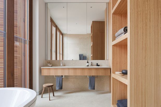 A bright, refined bathroom featuring a wall-mounted timber vanity with twin basins, a large mirrored wall, warm wood built-in shelving, pale stone or tile wall and floor finishes, and a partially visible freestanding white bathtub. Vertical windows with timber frames provide natural light. A small stool and towels add functional detail.