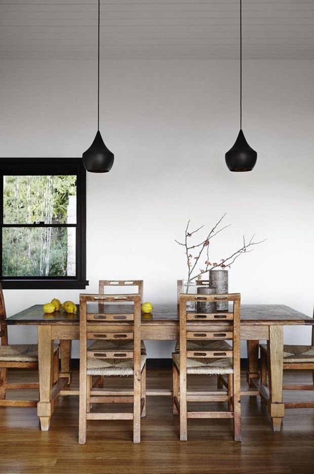 A dining room with a long rustic timber dining table and matching wooden bench seating and chairs. The room has white walls, dark timber-framed windows, hardwood flooring, and two suspended matte-black pendant lights. Simple natural branch arrangements and fruit sit on the table, creating a warm, understated setting.