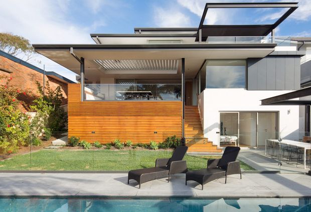 Contemporary multi-level residential home viewed from the backyard, featuring a flat roof with projecting glazed canopy, extensive outdoor living areas, timber-clad privacy screens, white rendered walls, and large glazed openings. A swimming pool and two outdoor lounge chairs occupy the foreground, with landscaped garden beds surrounding the house.