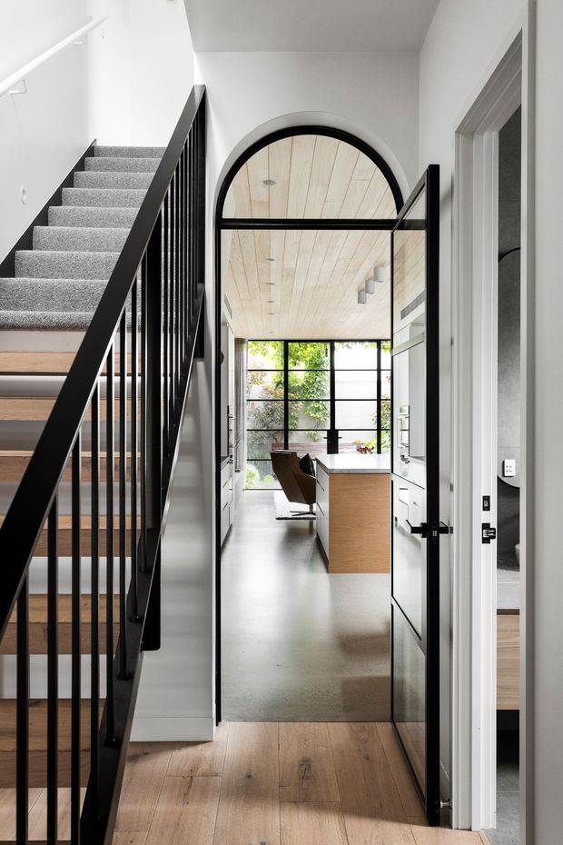 A bright contemporary interior view from a hallway or landing, showing a timber staircase with slim black metal balustrade, pale timber flooring, and a distinctive arched black-framed glazed door leading into a kitchen and dining area. The kitchen has white cabinetry, a timber island, polished concrete flooring, and expansive black-framed glazing. A pale timber-lined ceiling and minimalist linear lighting add warmth and architectural character.