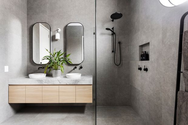 A contemporary bathroom with a floating light-wood double vanity, white vessel basins, rounded rectangular mirrors, grey stone-look tiled walls and floor, and a frameless glass shower enclosure with matte black fixtures. A small indoor plant adds greenery.