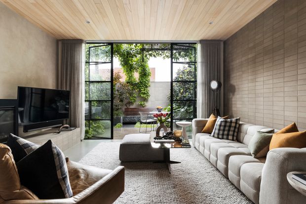 Contemporary living room with a large sectional sofa, leather armchairs, television and coffee table. The room features full-height black-framed glazing opening to a leafy courtyard, a warm timber-lined ceiling, textured neutral walls, and a large area rug. The palette is predominantly warm neutral with dark accents and abundant natural light.