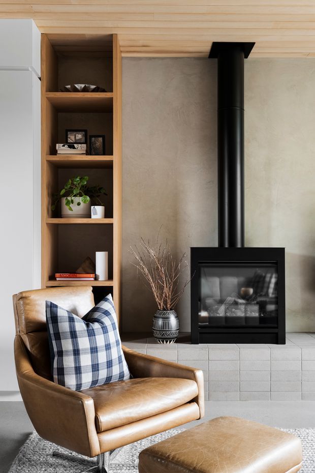 A warm, refined living room centred on a black wood-burning fireplace with a tall flue, set against a pale textured plaster wall and light grey tiled hearth. Built-in open timber shelving displays small decor and a plant, while a caramel leather armchair and matching ottoman with a blue-and-white checked cushion sit in the foreground. The ceiling and joinery use light timber, creating a contemporary rustic atmosphere.