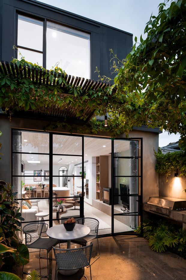 A contemporary two-storey home viewed from a leafy outdoor courtyard or terrace. Black-framed floor-to-ceiling glazed doors open to a bright living and dining interior, while a timber pergola covered in climbing plants creates a lush canopy overhead. The outdoor area includes a round dining table with woven black chairs, built-in shelving and a compact outdoor barbecue or kitchen zone against a rendered wall. Dark metal cladding, extensive glazing and warm interior lighting create a refined indoor-outdoor connection.