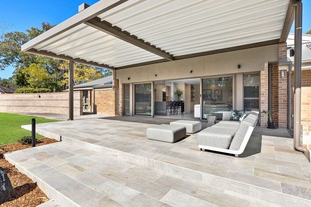 A bright, contemporary covered outdoor terrace adjoining a brick-clad house. The large pergola has a white slatted roof supported by dark metal beams and posts, while the terrace is finished in large pale stone-look tiles with broad steps leading to a lawn. Outdoor lounge furniture sits in front of glazed doors opening to the interior, with a dining area visible inside.