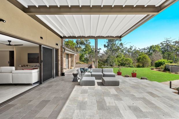 A spacious covered outdoor entertaining terrace with a motorised louvred roof, large-format grey stone-look paving, and contemporary outdoor seating. It opens directly to a bright living room through large sliding glass doors, with a landscaped lawn and trees beyond.