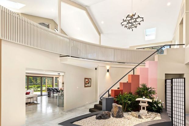 Bright, spacious contemporary interior featuring a dramatic double-height stair hall, sculptural black-and-pink open staircase, upper-level balustrade, skylights, and an indoor Japanese-inspired rock garden with plants and a small lantern. An adjoining dining or living area is visible through the opening at left.
