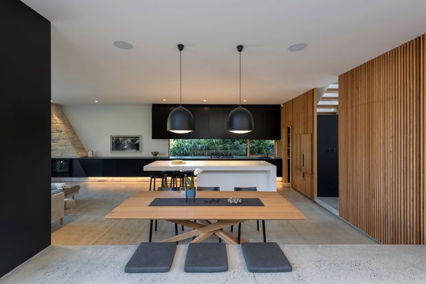 Open-plan contemporary kitchen and dining area viewed from a foreground island or bench. The space features a long light-wood dining table, black sculptural pendant lights, a white kitchen island with seating, dark cabinetry, warm timber slatted wall cladding, polished concrete flooring, and a large horizontal window above the kitchen bench. A stone-clad feature wall and integrated lighting add texture and warmth.